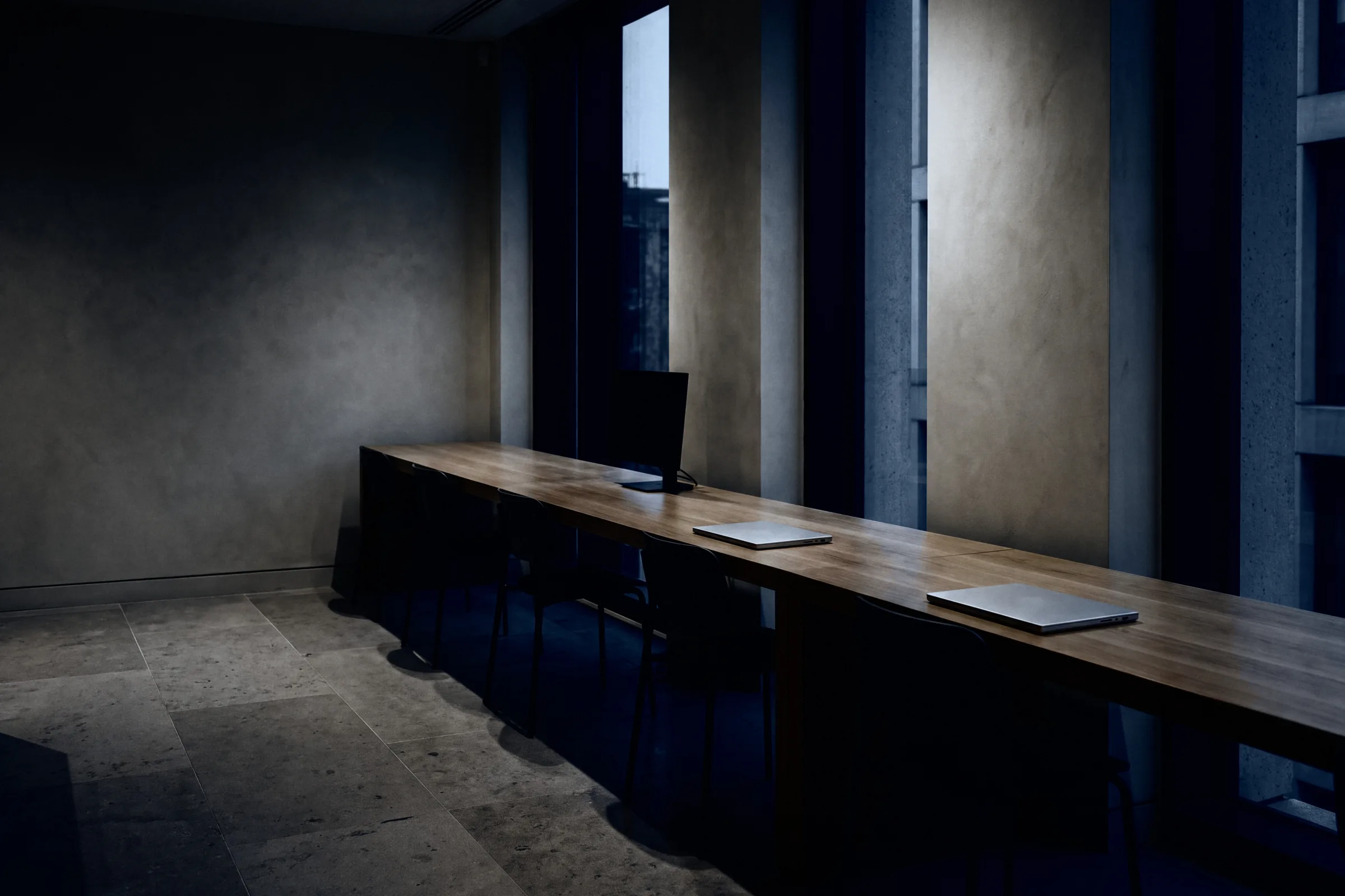 A dim room with one long wooden desk along the wall, two laptops on it