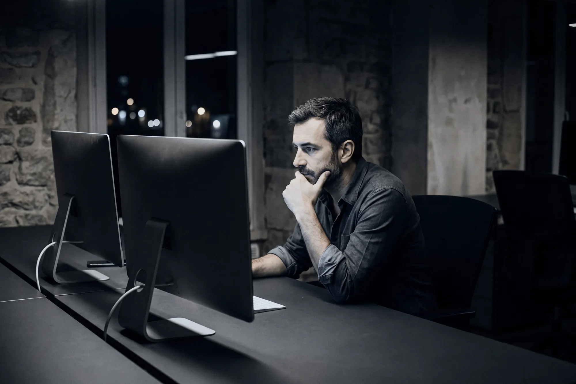 A trader at his desk at night, chin in hand, in front of two screens
