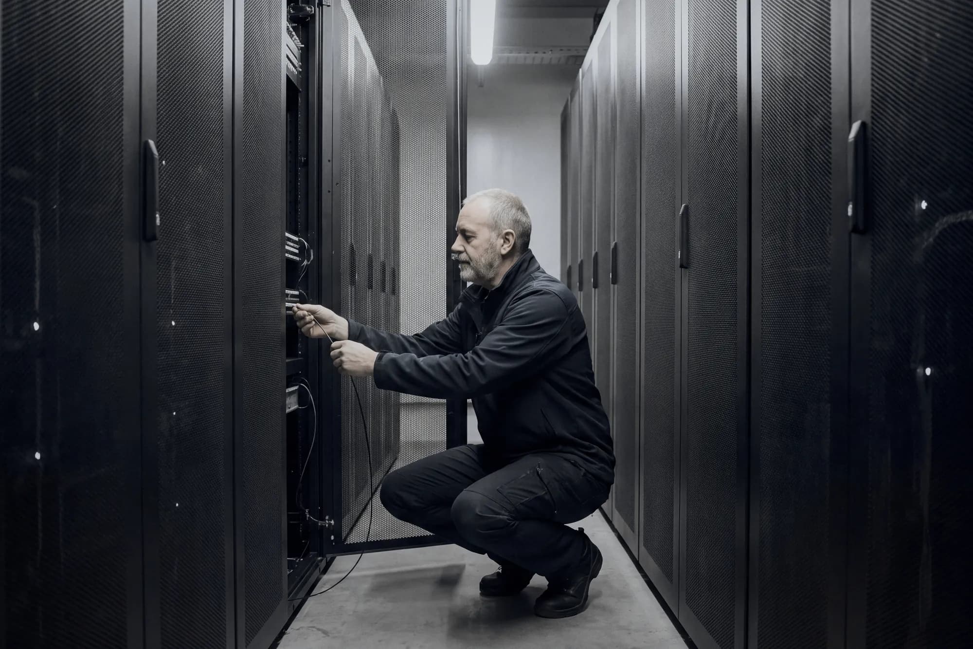 An engineer crouched at an open server rack, checking a cable